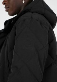Černá prošívaná péřová bunda s kapucí, vyrobená z texturované tkaniny, s diagonálním prošíváním a decentním logem na rukávu.