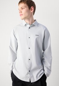 Lys grå langermet skjorte med knappdesign, sorte knapper og en liten svart "BOSS" logo på brystet. Glatt stoffstruktur.
