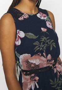 Blommig klänning i marinblått med rosa och gröna mönster. Har en halslinning med spetsdetaljer och ett tunt svart bälte runt midjan.