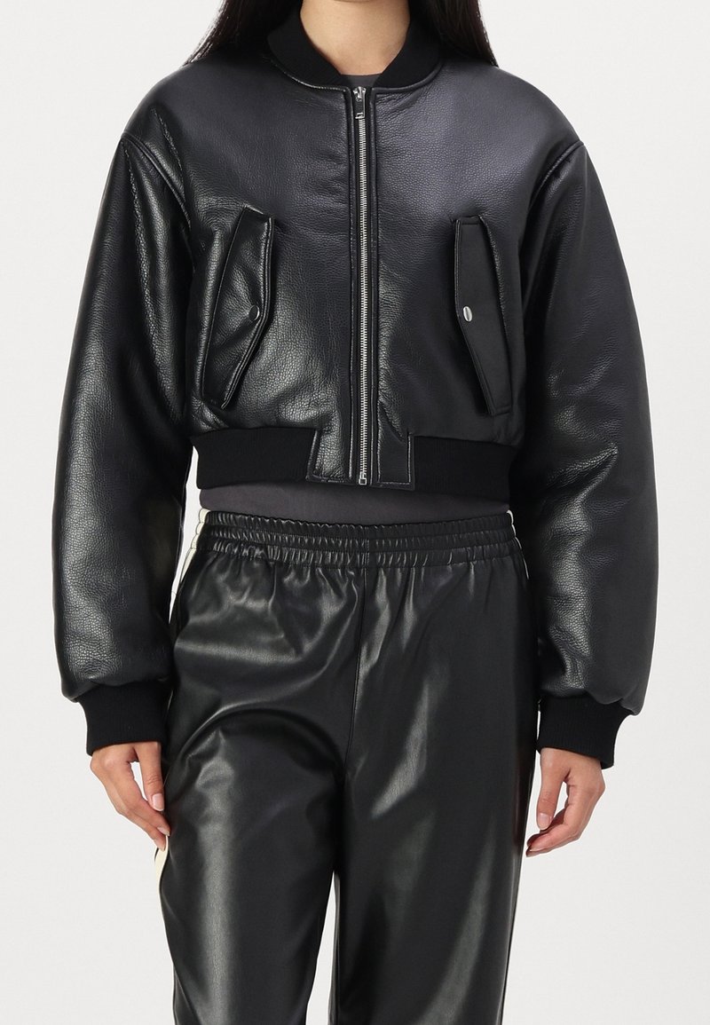 Femme portant une veste bomber en cuir noire courte avec fermeture éclair à l'avant et poches à rabat à pressions, associée à un pantalon en cuir noir.