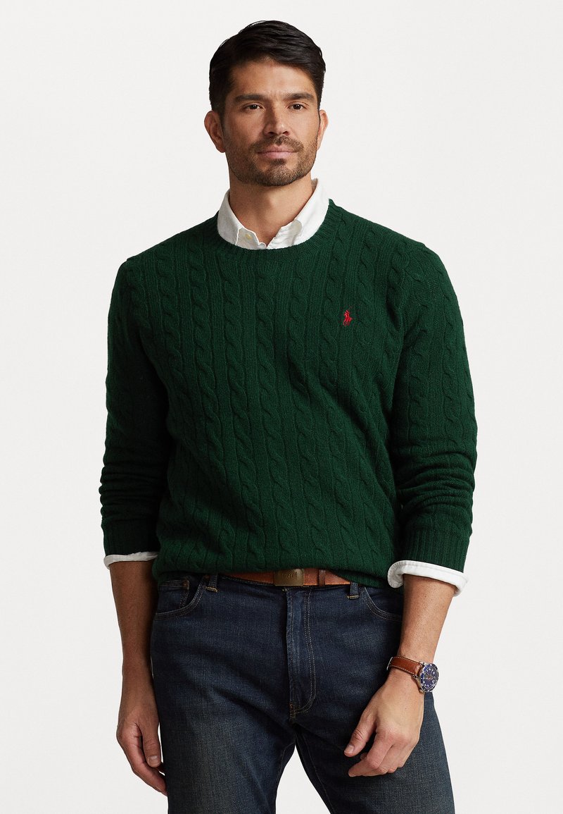 Suéter verde de punto trenzado, de cuello redondo, con un pequeño logotipo rojo en el pecho. Llevado sobre una camisa blanca con cuello, combinado con jeans oscuros y un cinturón marrón.