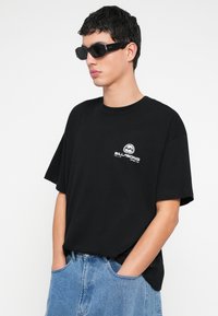 Czarny bawełniany t-shirt z krótkimi rękawami, z małym białym logo Billabong na piersi. Dopasowany do jasnoniebieskich spodni dżinsowych.