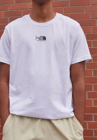 Camiseta de algodón blanca con un pequeño logo negro que dice "THE NORTH FACE"; escote redondeado, mangas cortas, corte holgado, textura suave.