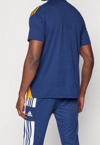 Homme portant une tenue de sport bleue avec des accents jaunes, comprenant un t-shirt à manches courtes et un pantalon de survêtement Adidas à rayures blanches.