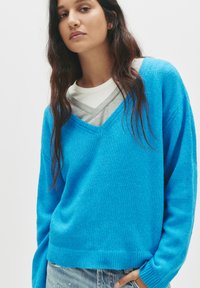 Blauer V-Ausschnitt-Pullover aus weichem Strickmaterial, mit langen Ärmeln und einem lässigen Schnitt. Darunter ist ein graues Shirt getragen.