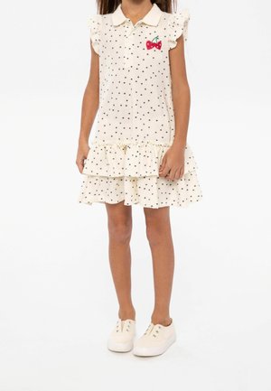 Robe crème à pois bleu marine, manches courtes à volants, col et broderie de cerises. Volants à étages à l'ourlet. Associée à des baskets blanches.