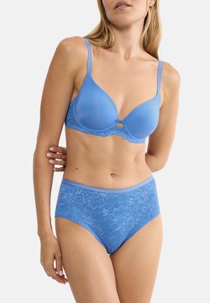 Frau trägt passenden blauen BH mit glatten Cups und hoch taillierte Pantys aus floraler Spitze vor einfachem Hintergrund.
