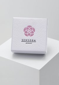 Bela teksturirana škatlica za nakit z rožnatim cvetličnim logotipom in napisom "SENSORA JEWELRY" v črni barvi. Oblika škatlice je kvadratna in kompaktna.