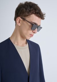 Blazer navy con colletto sciallato, indossato sopra una maglietta beige a girocollo. Occhiali da sole scuri con montatura rettangolare. Texture del tessuto liscia.