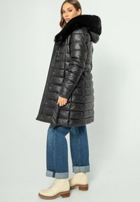 Zwarte pufferjas met een nepbont kraag, voorzien van een rits- en drukknoopsluiting, gequilt textuur en een knielange ontwerp. Draagt over blauwe spijkerbroeken.