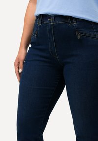 Tummansinisiä farkkuhousuja, joissa on istuva malli, lievä venymä, etutaskut vetoketjulla ja näkyvät ompeluseikat.