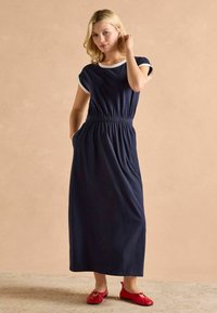 Námornícka modrá maxi šaty s krátkymi rukávmi, bielym lemovaním, elastickým pásom a bočnými vreckami. Vypárované s červenými plochými topánkami.