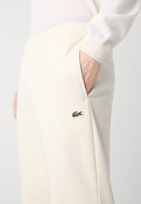 Crème-kleurige joggers gemaakt van zachte stof met een relaxte pasvorm, voorzien van een geborduurd groen krokodil-logo en zijzakken.