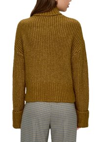 Senfgelber Strickpullover mit hohem Kragen, gerippten Bündchen und cropped Saum. Die Textur ist grob mit einem sichtbaren Strickmuster.