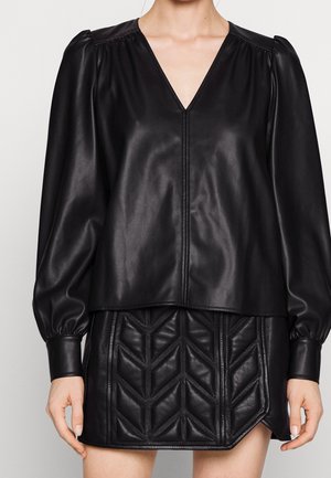 Kvinde iført en sort bluse i kunstlæder med lange ærmer og V-hals samt en sort quiltet mininederdel med geometriske mønstre.