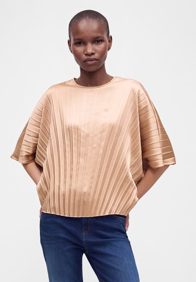 Femme aux cheveux courts portant un haut beige brillant plissé et un jean bleu, debout les mains dans les poches devant un fond uni.