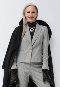 Grau kariertes Anzug mit einem taillierten Blazer und passender Hose, kombiniert mit einem schwarzen Rollkragenpullover und schwarzen Lederhandschuhen. Silberne Halskette als Akzent.