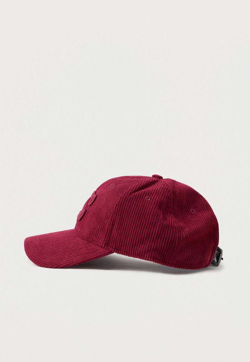 Gorra de béisbol de pana roja con visera curva, que cuenta con seis agujeros de ventilación, una correa de tela en la parte trasera y una superficie texturada.