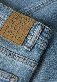 Denimjeans med blå vask, med et teksturert brunt merke. Merkets tekst lyder "FORWARD JEANS WEAR SINCE 1986." Dobbeltsydde sømmer.