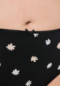 Sous-vêtements en coton noir avec un motif floral en blanc et beige clair, arborant un petit nœud noir en accent à la taille.
