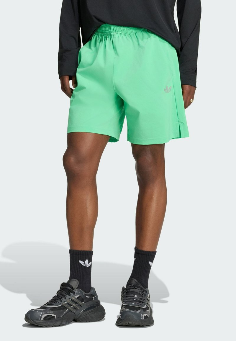 Person trägt leuchtend grüne Shorts, schwarzes Langarmshirt, schwarze Adidas-Socken und schwarze Sportschuhe und steht vor einem weißen Hintergrund.