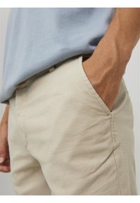 Beige texturované kalhoty s volným střihem, mají přední kapsu a viditelný pas, spárované se světle modrou košilí.