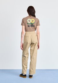 T-shirt marrone con grafiche di girasoli e la parola "DICKIES" sul retro, abbinato a pantaloni beige a vita alta e scarpe nere.