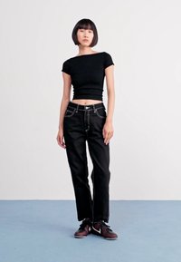 Top curto preto, jeans pretos com costura branca, em pé num chão azul claro. Calçado são ténis casuais escuros. Fundo branco liso.