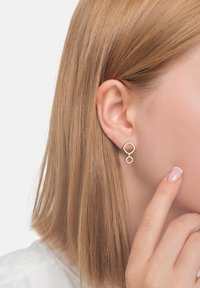 Guldörhängen med geometrisk design har två sammanflätade former: en oval ovanför en kvadrat. Modellen har rakt, axellångt hår.