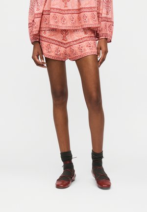 Personne portant un short rouge à motifs et un haut assorti, des chaussettes foncées et des ballerines rouges, se tenant devant un fond clair uni.