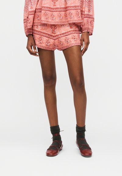 Personne portant un short rouge à motifs et un haut assorti, des chaussettes foncées et des ballerines rouges, se tenant devant un fond clair uni.