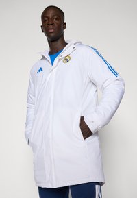 White insulated parka with a hood, blue accents, and Real Madrid logo. Features side pockets and a buttoned front closure.