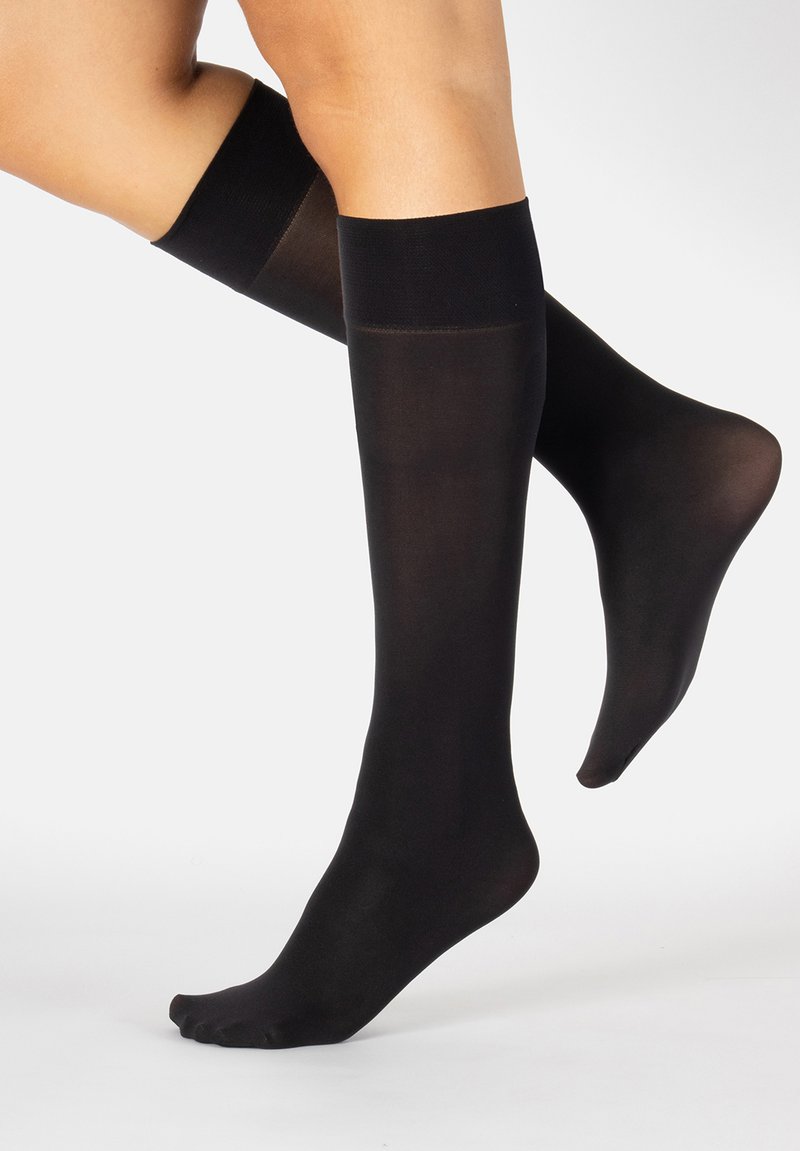Piernas vistiendo medias de nylon negras transparentes hasta la rodilla con bandas elásticas en la parte superior, un pie en punta, contra un fondo blanco liso.