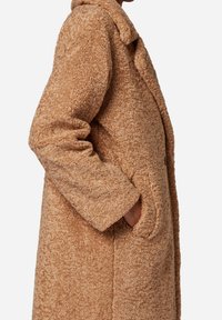 Beige pluizige jas met een gestructureerde, zachte stof. Voorzien van een kraag, zijzakken en een ontspannen pasvorm. Naden zijn zichtbaar langs de randen.