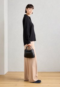 Bolsa de mão preta, tecida, com uma forma estruturada, segurada por uma figura que veste uma camisa preta de mangas longas e calças largas bege.