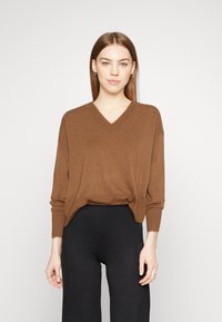 Bruine V-hals trui met lange mouwen, geribde manchetten en zoom, gedragen over zwarte broeken. Zachte textuur zonder zichtbare patronen of versieringen.