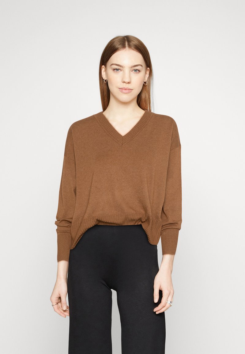 Bruine V-hals trui met lange mouwen, geribde manchetten en zoom, gedragen over zwarte broeken. Zachte textuur zonder zichtbare patronen of versieringen.