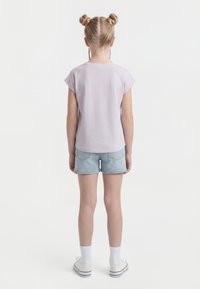 Mlada deklica stoji obrnjena stran, oblečena v svetlo vijolično majico, jeansa kratke hlače, bele nogavice in bele superge, z lasmi v dveh figah.