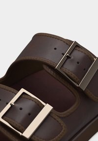 Sandale en cuir marron avec deux boucles argentées ajustables et couture visible le long des brides.