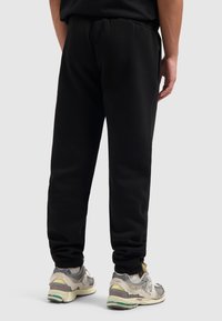 Zwarte trainingsbroek met een elastische tailleband en manchetrokken pijpen. Draagt een paar grijze en gele sneakers. Het materiaal lijkt zacht en textuur te hebben.