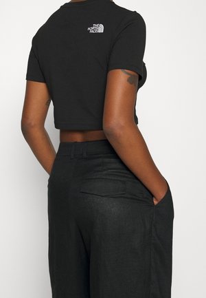 Black cropped t-shirt with short sleeves and a small white logo on the back, paired with high-waisted dark trousers featuring back pockets.