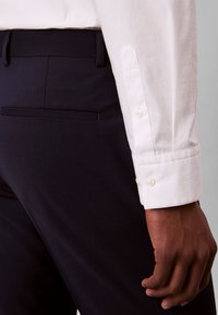 Persona che indossa pantaloni neri su misura e una camicia bianca a maniche lunghe con polsini abbottonati, braccio appoggiato di fianco.