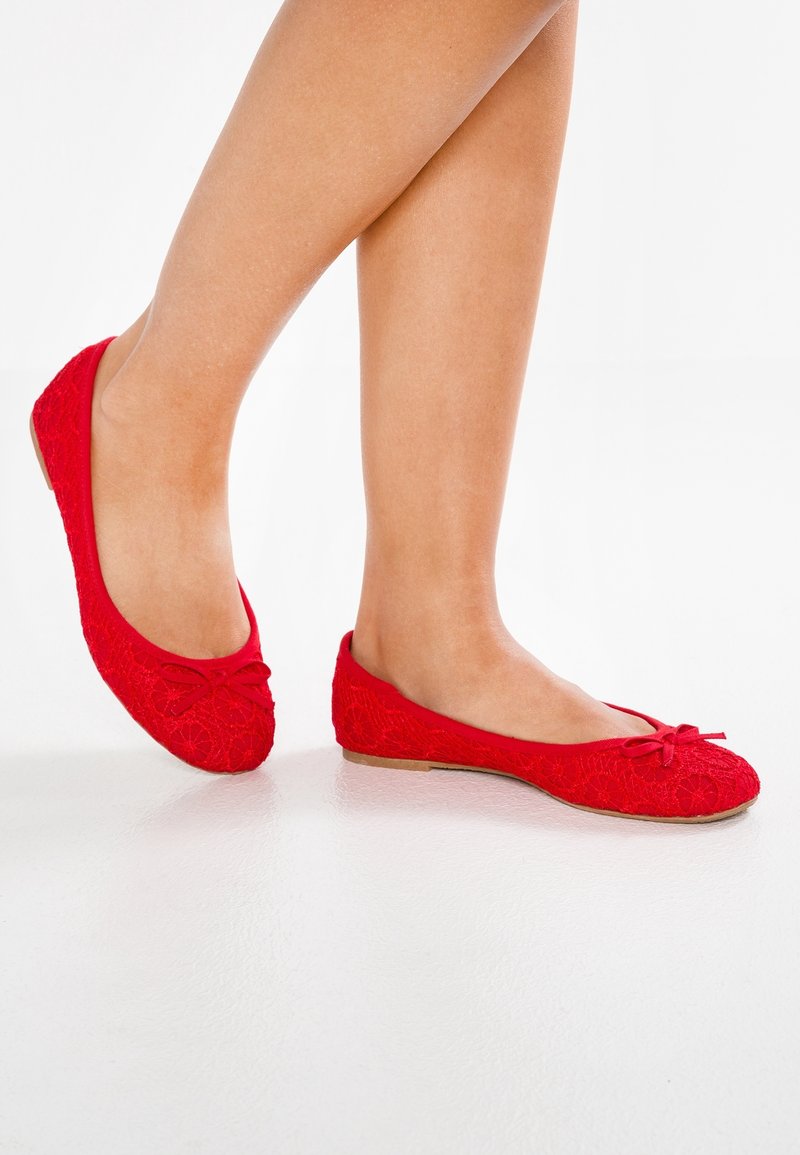 Gambe che indossano scarpe da balletto rosse in pizzo con piccoli fiocchi, in piedi su una superficie bianca.