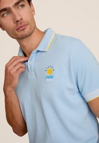 Lichtblauwe polo met korte mouwen, met een kraag met gele rand, geborduurd zon- en golfontwerp en een gestructureerde stof.