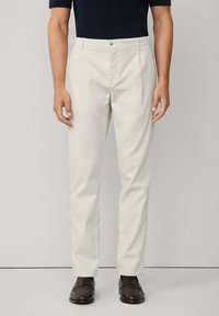 Muškarac u bež ravnim hlačama, tamnosmeškim kožnim loafericama i tamnoplavoj košulji kratkih rukava na neutralnoj pozadini.