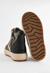 Sneakers alte in materiali verde oliva e nero, con lacci arancioni, zip laterale e suola in gomma testurizzata con motivo a zigzag.