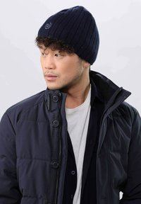Navy ribbed knit beanie with a logo, worn with a dark puffer jacket and white shirt, showcasing texture and layered design details.