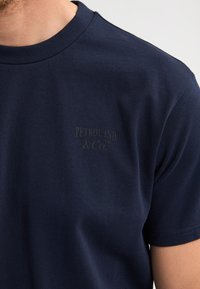 Marineblauw katoenen T-shirt met korte mouwen, met een subtiel gedrukt logo "PETROL IND & Co" op de linkerborst. Soepele textuur.