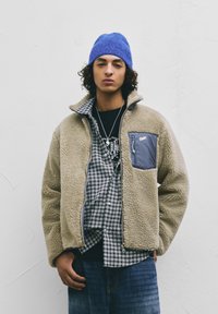 Mladostna oseba s kodrastimi lasmi, ki nosi modro kapo, bež flis jakno z zadrgo, karo srajco, črno majico z grafikami in modre kavbojke.