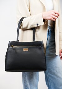 Zwarte leren tote bag met een gestructureerde afwerking, gouden ritsaccent en een rechthoekige vorm, vastgehouden door een persoon in een roomkleurige blazer.